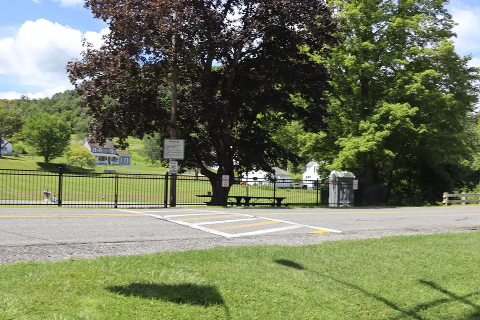 A crosswalk leads from the grassy parking area to Tyringham Town Park, which is accessible through a latched metal gate. A portable restroom can be seen in the background.