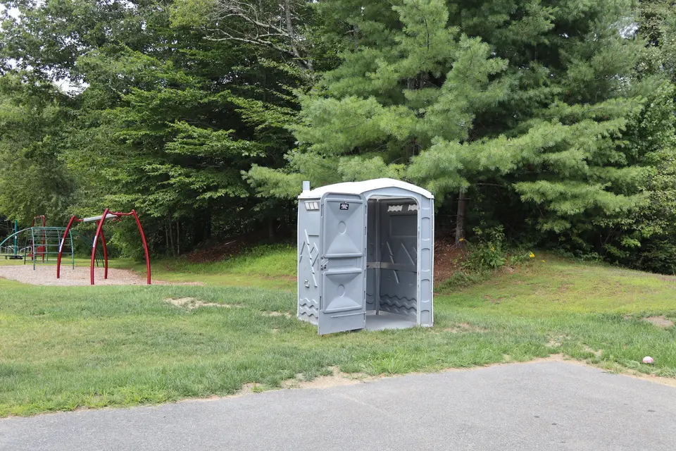 A portable restroom located in a corner of the Town Hall parking lot, in front of a playground area.